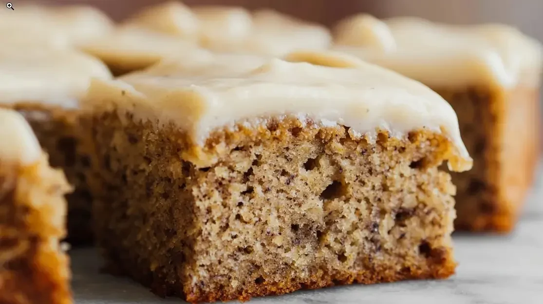 The Easiest & Most Delicious Banana Cake with Cake Mix 1 Close-up of a moist banana cake square topped with creamy vanilla frosting on a marble surface.