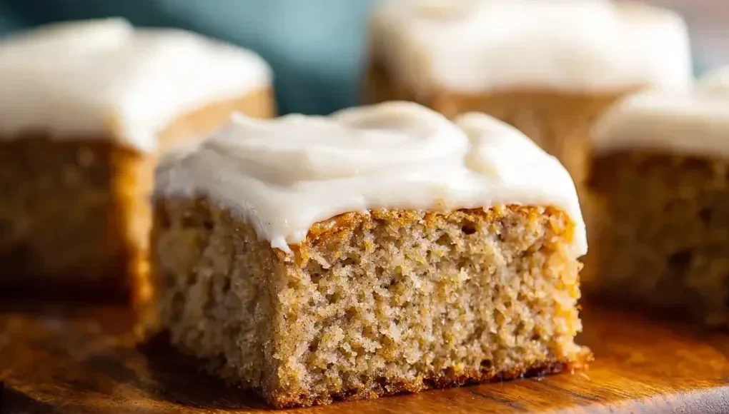 Square piece of moist banana cake with vanilla frosting on a wooden serving board