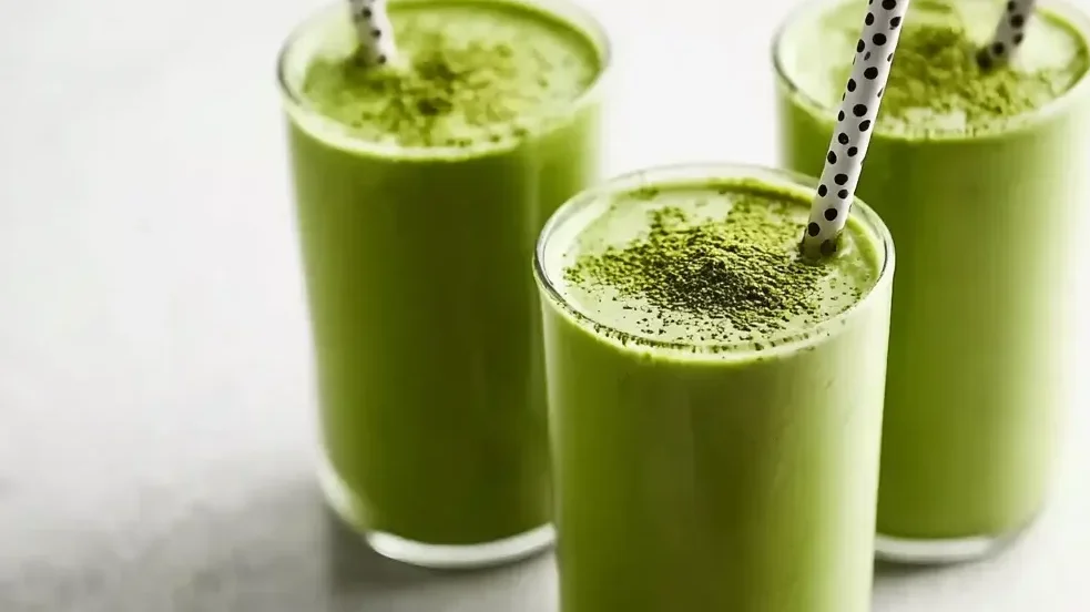 Three glasses of creamy matcha protein shake with black-and-white polka dot straws on a light background, sprinkled with matcha powder