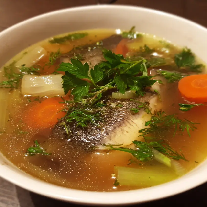 A close-up of a warm, inviting bowl of clear fish bone broth soup with chunks of fish, carrots, onions, celery, and fresh herbs.