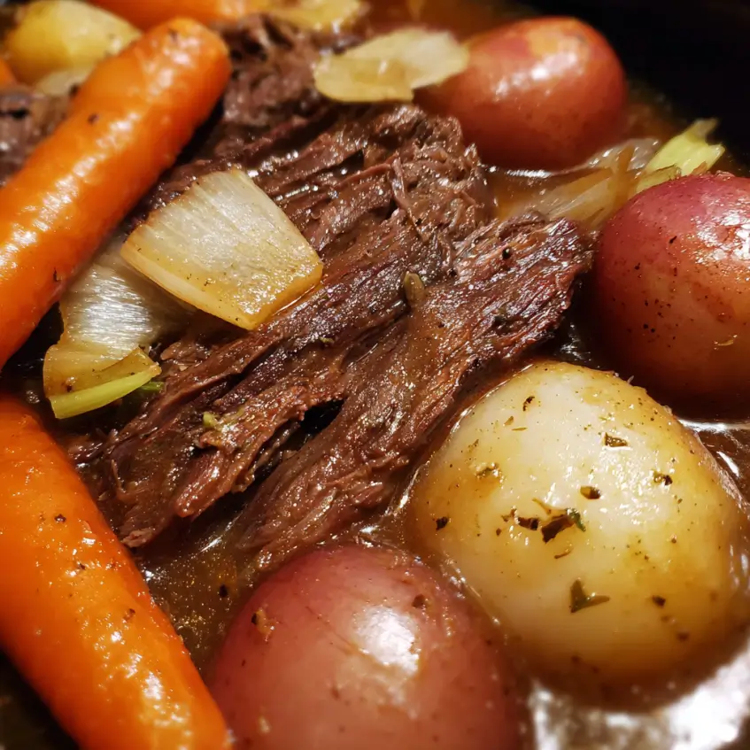 A close-up of a hearty beef roast crockpot recipe, featuring tender shredded beef, vibrant carrots, potatoes, and onions in a rich, dark brown gravy.