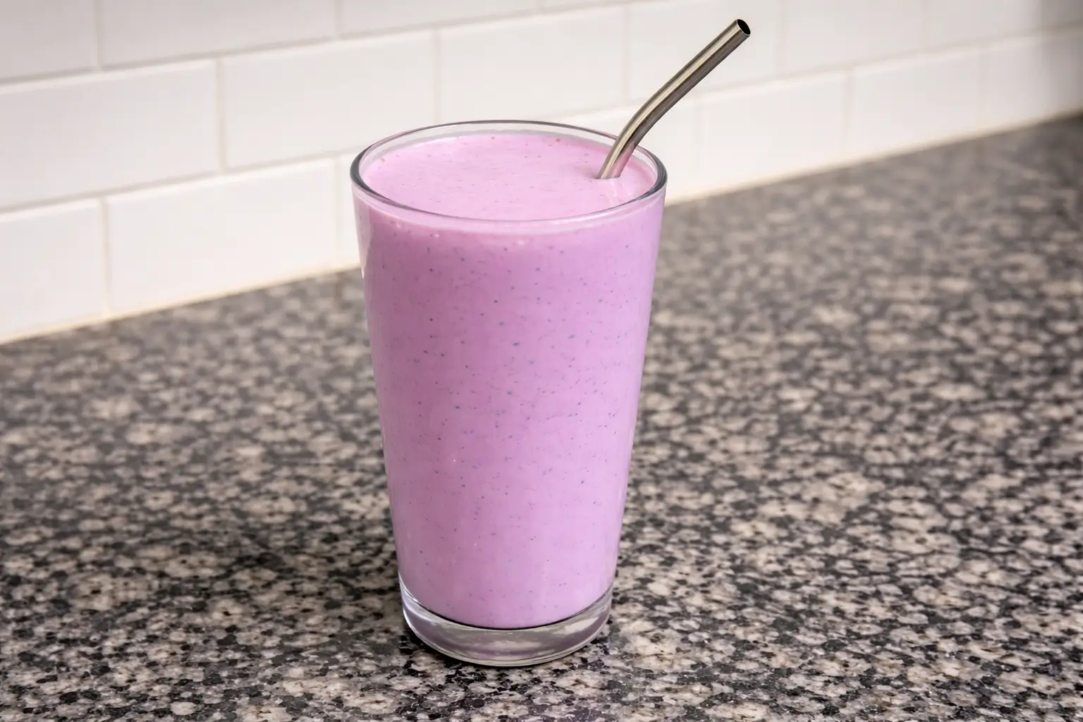 Ultra-creamy blueberry spinach smoothie in clear glass with stainless steel straw on dark marble countertop, vibrant purple-blue blend with subtle green spinach flecks, creamy yogurt texture, fresh healthy breakfast drink